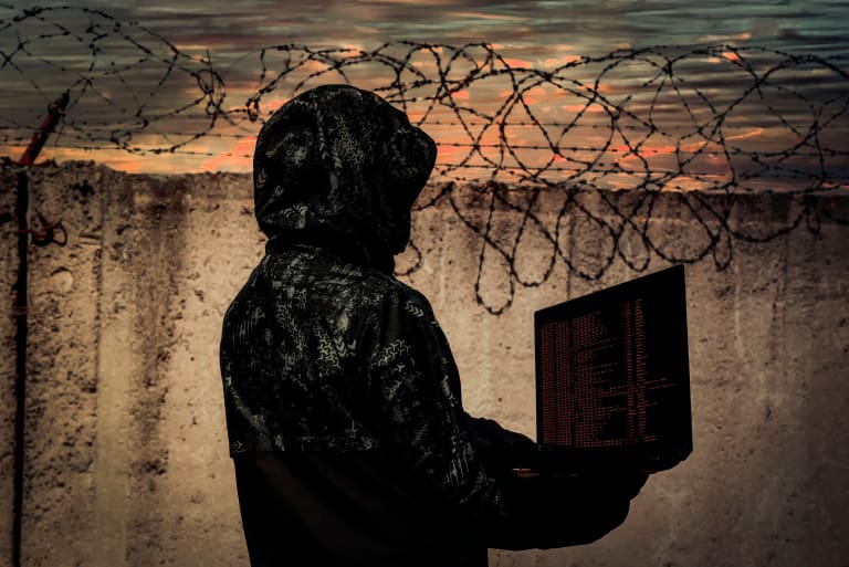 A man in a hoodie holding a laptop in front of barbed wire.