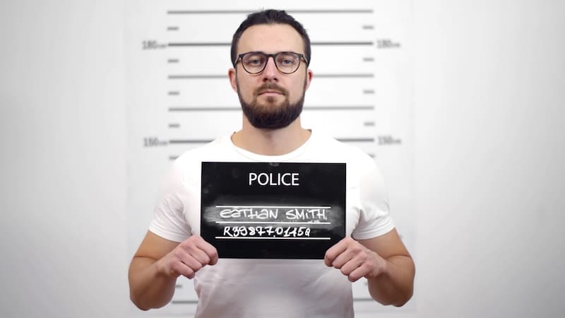 Mugshot of young guilty bearded man at police station. Handsome criminal guy in glasses holding signboard posing for mug shot with height chart on background.