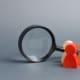 A magnifying glass focusing on two wooden figurines, one painted red and the other in natural wood, set against a plain grey background.
