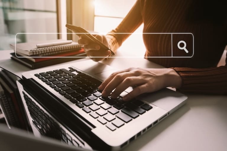A person uses a laptop while looking at their phone. A search bar is overlayed on the image.